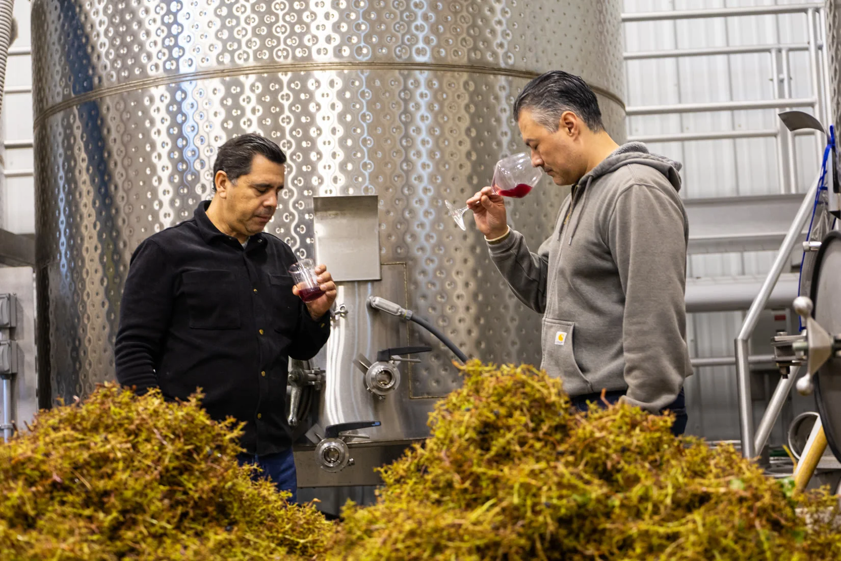 Two winemakers tasting red wine between steel tanks
